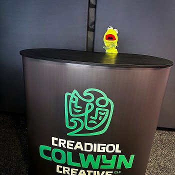 Pyped yn ymddangos tu ol i arwydd gyda logo Creadigol Colwyn Creative